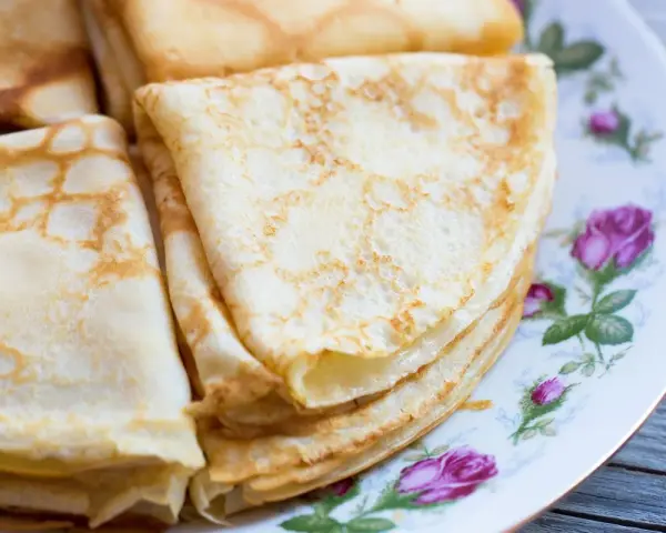 Deliziose crepes senza latte, piegate e impilate su un piatto decorato con rose. Perfette per una colazione leggera o uno spuntino.