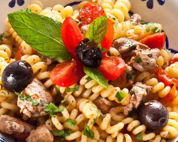 Pasta con pesce spada, pomodorini, olive nere e menta fresca in una ciotola decorata. Un piatto estivo che profuma di mare.