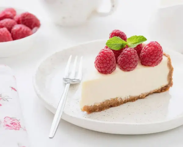 Una fetta di torta fredda allo yogurt guarnita con lamponi freschi e menta, su un piatto bianco con una forchetta.
