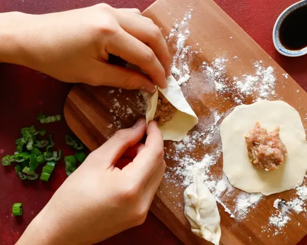 Mani che preparano il ripieno per ravioli su un tagliere di legno, con erba cipollina e salsa di soia a lato.