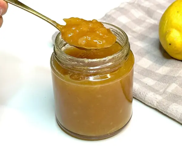 Un cucchiaio d'oro raccoglie la densa marmellata di limoni da un barattolo di vetro. Due limoni freschi riposano su un tovagliolo a quadretti.