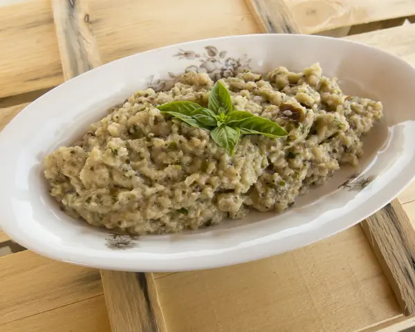 Crema di melanzane rustica, guarnita con basilico fresco, servita in un piatto ovale su un tavolo di legno grezzo.
