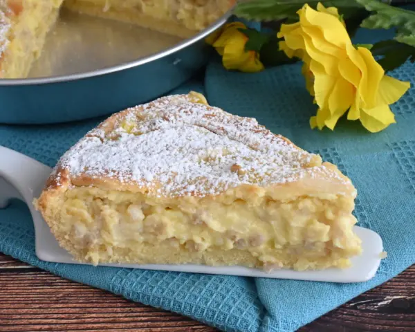 Una fetta di pastiera napoletana con crema pasticcera, cosparsa di zucchero a velo, su un piatto bianco, con fiori gialli sullo sfondo.