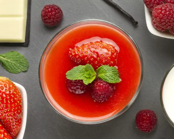 Un bicchiere di coulis di fragole con fragola, lamponi e menta. Accanto, fragole fresche, cioccolato bianco, lamponi e un bicchiere di latte.