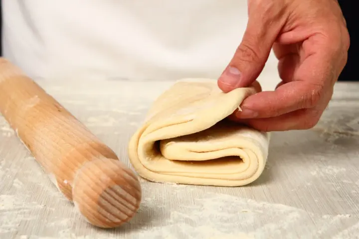 Mani che piegano un impasto per pasta fatta in casa, con un mattarello di legno accanto, cosparso di farina.