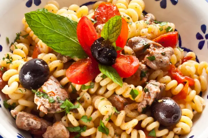 Pasta con pesce spada, pomodorini, olive nere e menta fresca in una ciotola decorata. Un piatto estivo che profuma di mare.