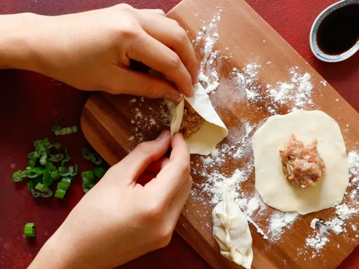 Mani che preparano il ripieno per ravioli su un tagliere di legno, con erba cipollina e salsa di soia a lato.