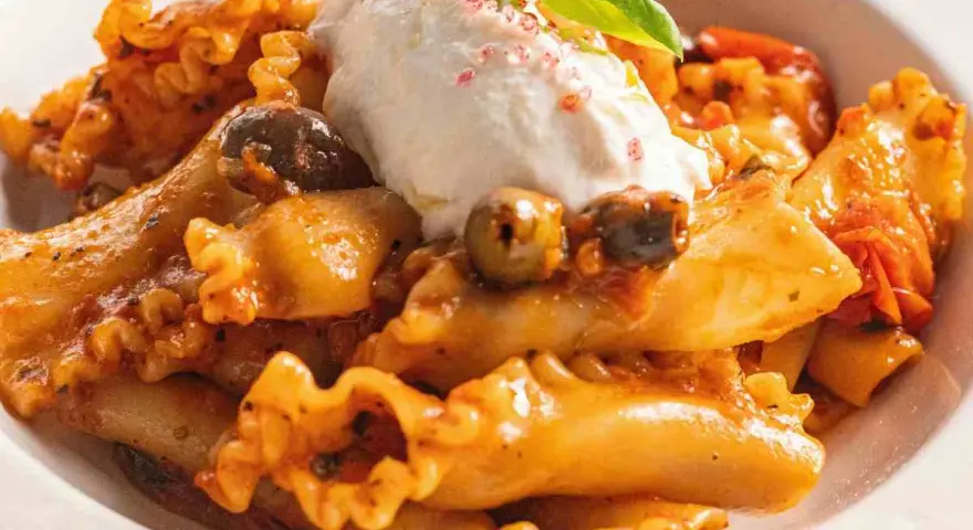 Pasta con melanzane, condita con un sugo ricco, olive e una generosa quenelle di ricotta fresca, guarnita con basilico.