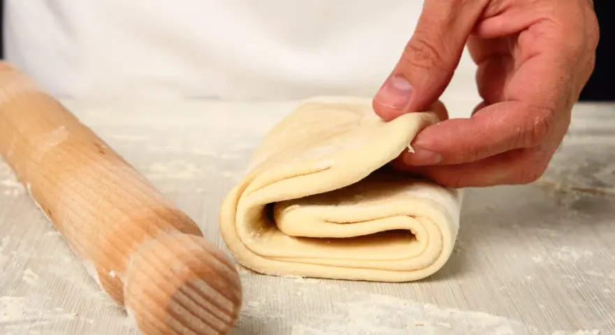 Mani che piegano un impasto per pasta fatta in casa, con un mattarello di legno accanto, cosparso di farina.