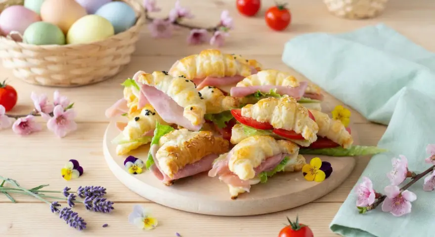 Delizioso antipasto di Pasqua con croissant farciti, uova colorate in un cesto e fiori primaverili.