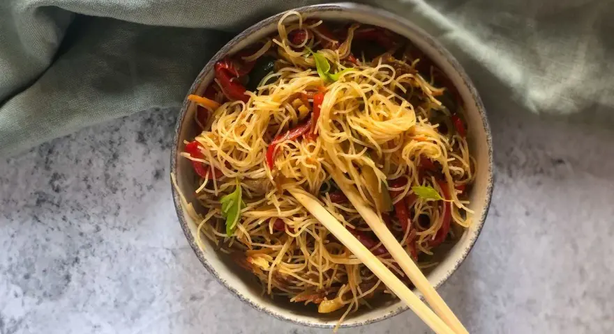 Un piatto invitante di spaghetti di riso con verdure colorate, come peperoni rossi e zucchine, guarnito con basilico fresco e servito con bacchette.