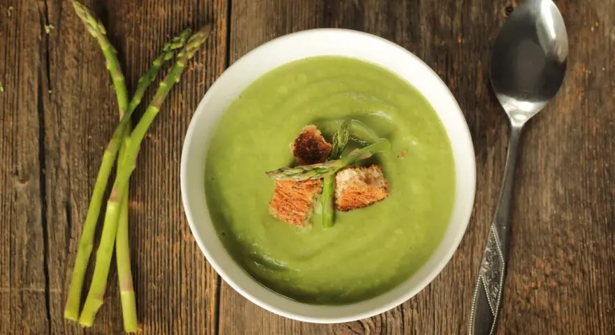 Una ciotola di cremosa crema di asparagi, guarnita con crostini e germogli freschi, accanto a un mazzo di asparagi su un tavolo di legno.