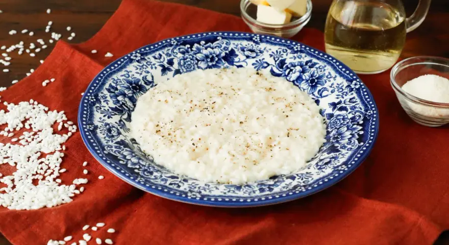 Un cremoso riso in bianco, condito con pepe nero, servito su un piatto decorato. Accanto, burro, olio e parmigiano grattugiato, ingredienti per un piatto semplice ma gustoso.