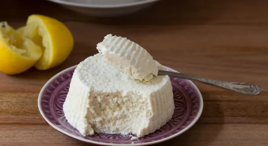 Ricotta fatta in casa su un piatto viola, con limone tagliato a metà sullo sfondo. Un cucchiaino preleva una porzione cremosa.