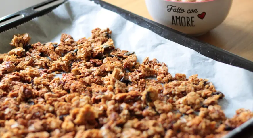 Granola fatta in casa appena sfornata su carta da forno, con una ciotola di frutta secca sullo sfondo.