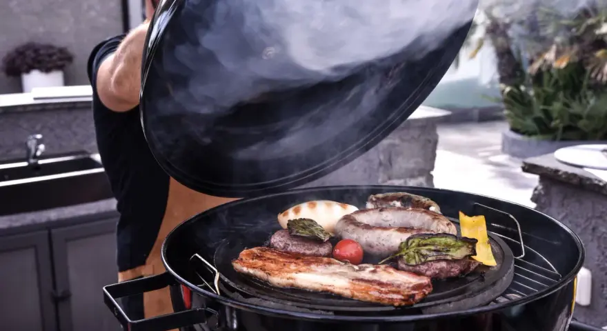Un uomo solleva il coperchio di una grigliata di carne fumante, rivelando salsicce, pancetta, hamburger e verdure.