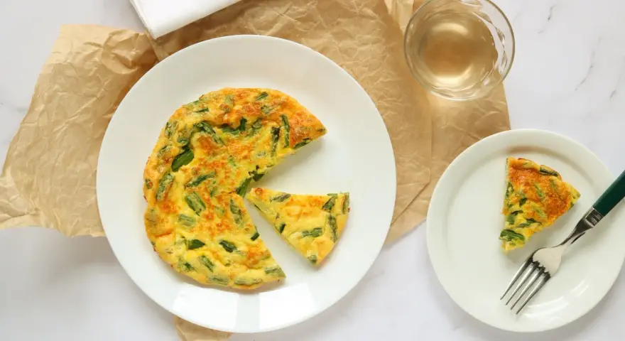 Deliziosa frittata di asparagi al forno, servita su un piatto bianco con una fetta a parte e un bicchiere di vino.