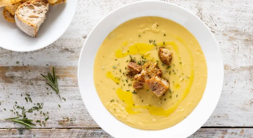Una cremosa vellutata di ceci, guarnita con crostini dorati, olio d'oliva e rosmarino fresco, su un tavolo di legno rustico.