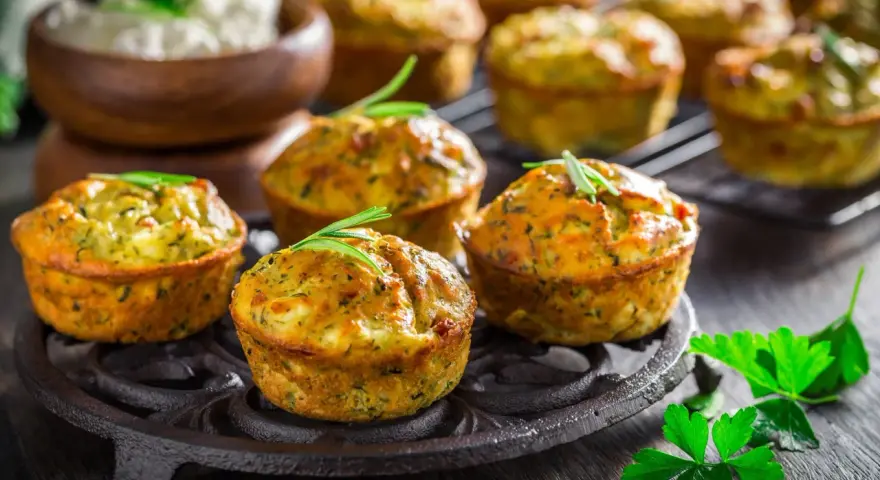 Tortini di zucchine dorati e fragranti, guarniti con rosmarino, su un piatto di ghisa. Sullo sfondo, una ciotola di formaggio fresco.