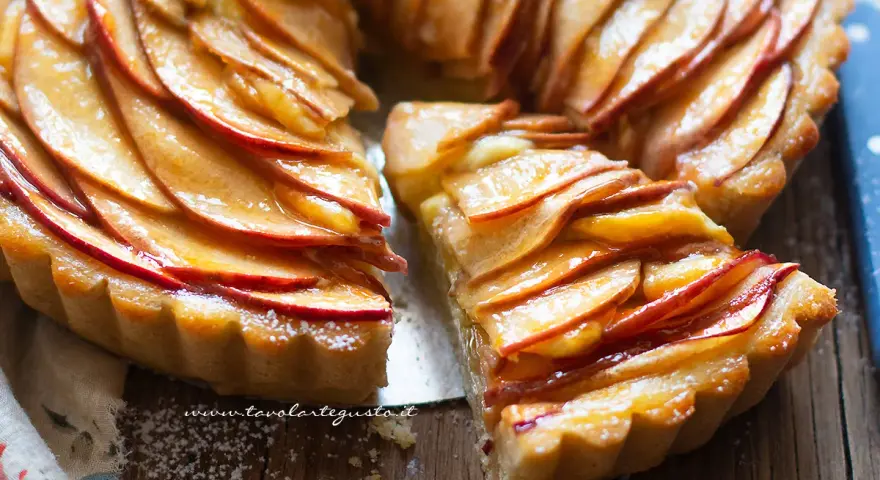 Una deliziosa crostata di mele, con fette di mela lucide disposte a spirale su una base dorata. Una fetta è stata tagliata, rivelando la morbidezza del ripieno.