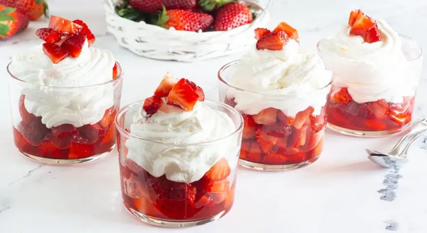 Coppette di fragole con panna montata e pezzetti di fragola fresca in cima. Un cestino di fragole sullo sfondo.