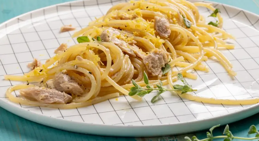 Un piatto di pasta tonno e limone, condita con scorza di limone grattugiata e erbe aromatiche fresche, servita su un piatto a scacchi bianchi.