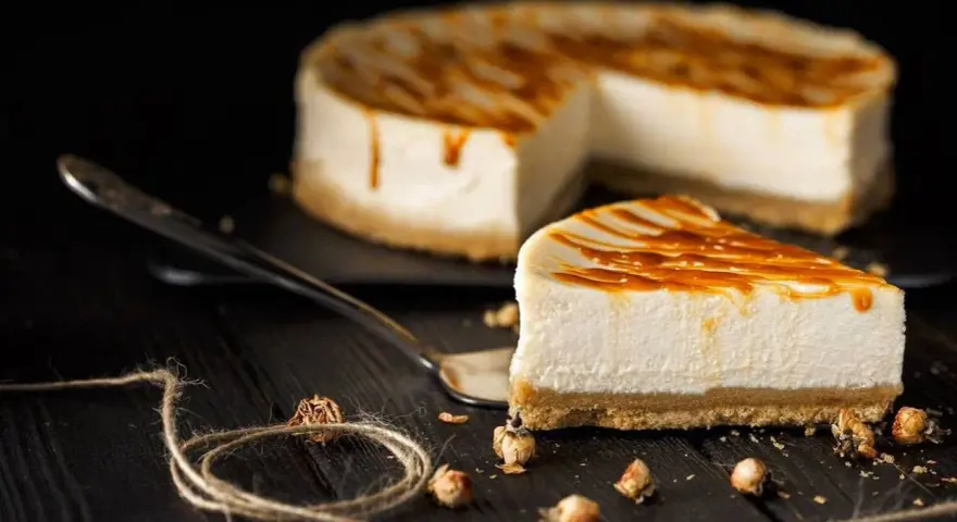 Una fetta di cheesecake senza panna, con glassa caramellata, su un piatto da portata. Sullo sfondo, la torta intera.