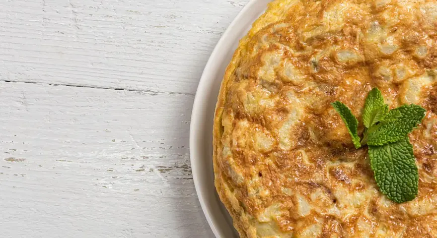 Frittata di cipolle dorata e soffice, guarnita con foglie di menta fresca, su un tavolo di legno bianco.