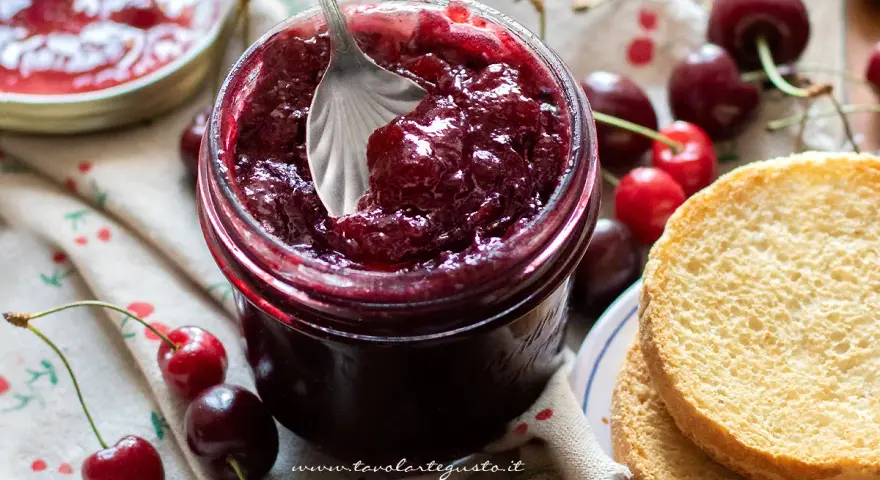 Un barattolo pieno di deliziosa marmellata di ciliegie, con ciliegie fresche e fette biscottate accanto.