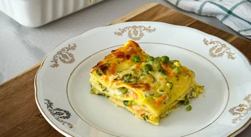 Una fetta di lasagne di verdure, con piselli e carote, su un piatto bianco decorato.