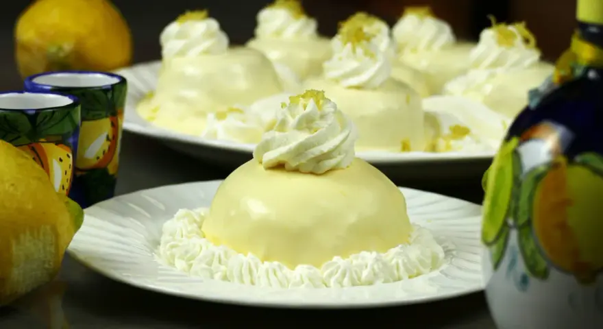 Dolcetti rotondi con glassa al limone e panna montata, guarniti con scorza di limone. Sullo sfondo, limoni freschi e bicchierini decorati.