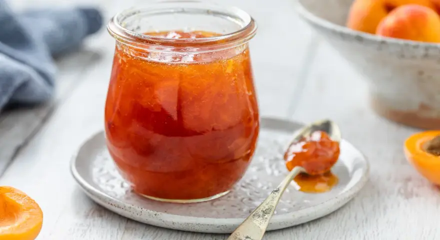 Vasetto di marmellata di albicocche, con un cucchiaino carico e albicocche fresche sullo sfondo.