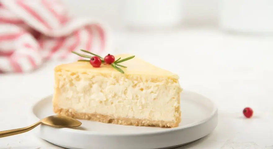 Una fetta di cheesecake yogurt greco, guarnita con bacche rosse e rosmarino, su un piatto bianco con un cucchiaio dorato.