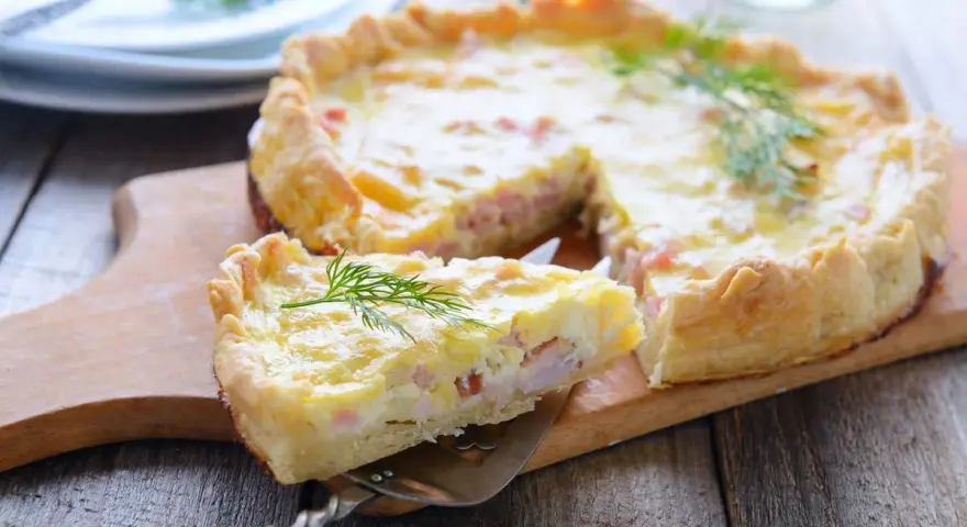 A slice of prosciutto and cheese tart is lifted from the whole tart on a wooden board, garnished with dill.