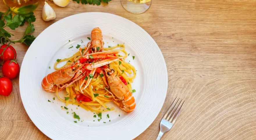 Un piatto di linguine agli scampi, condite con pomodorini e prezzemolo, accompagnato da ingredienti freschi e un bicchiere di vino bianco.