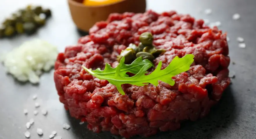 Battuta di fassona cruda, guarnita con capperi e rucola, con tuorlo d'uovo e cipolla tritata sullo sfondo.