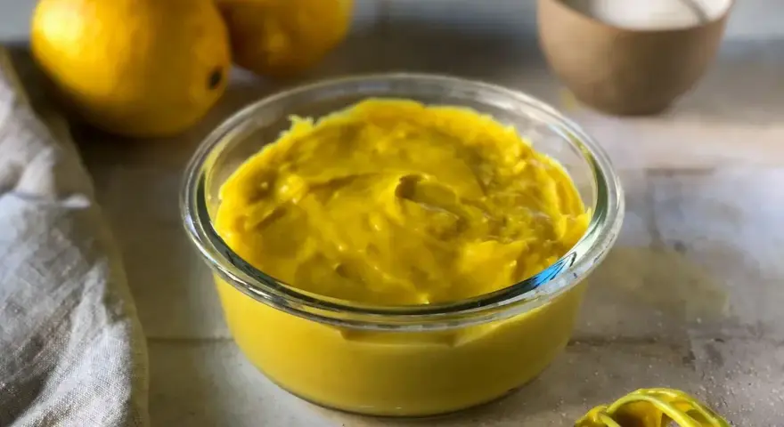 Crema pasticcera senza uova, densa e vellutata, in una ciotola di vetro. Limoni e zucchero sullo sfondo, pronti per un dolce delizioso.