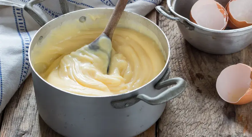 Crema pasticcera veloce in pentola, con cucchiaio di legno che mescola. Guscio d'uovo accanto, pronto per un dolce.