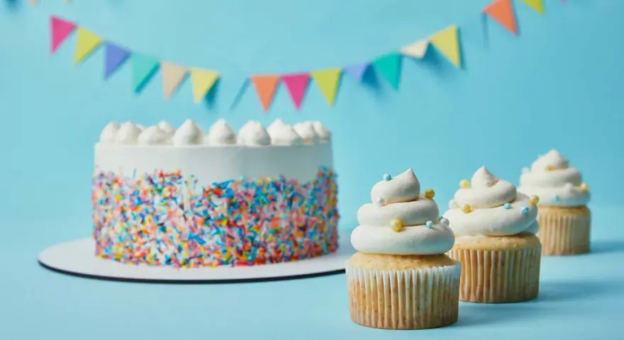 Torta bianca con codette colorate e cupcake decorati con soffice crema al burro, su sfondo azzurro con festoni.