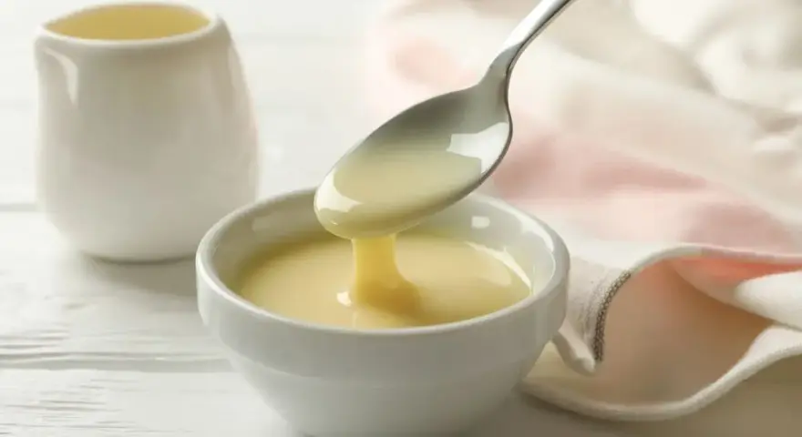 Un cucchiaio versa un denso latte condensato bimby in una ciotola bianca. Sullo sfondo, una piccola brocca e un tovagliolo rosa.