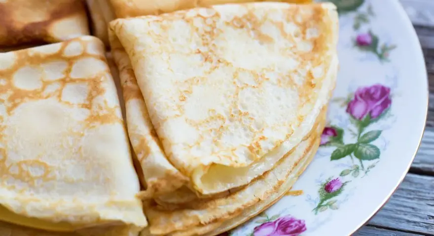 Deliziose crepes senza latte, piegate e impilate su un piatto decorato con rose. Perfette per una colazione leggera o uno spuntino.
