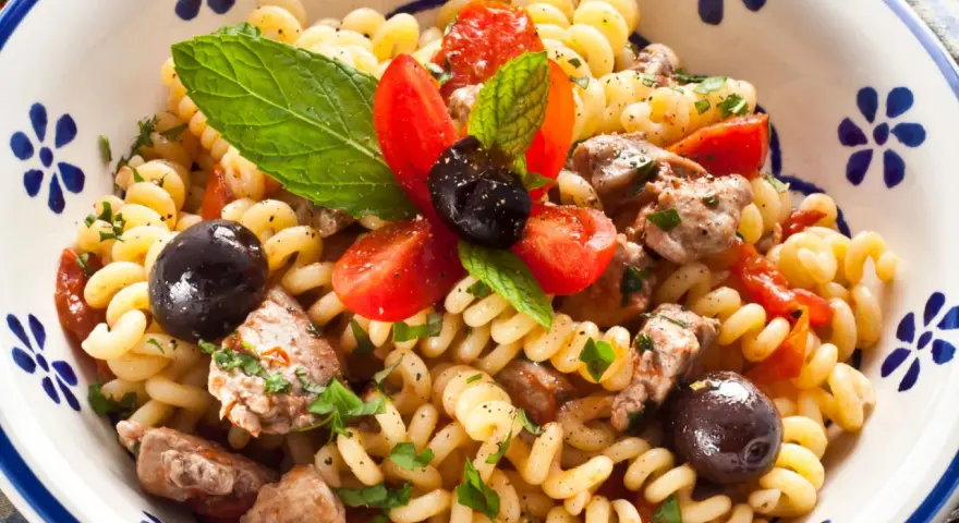 Pasta con pesce spada, pomodorini, olive nere e menta fresca in una ciotola decorata. Un piatto estivo che profuma di mare.