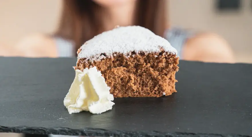 Una fetta di pan di spagna al cacao, soffice e spolverata di zucchero a velo, servita con panna montata su una lavagna scura.