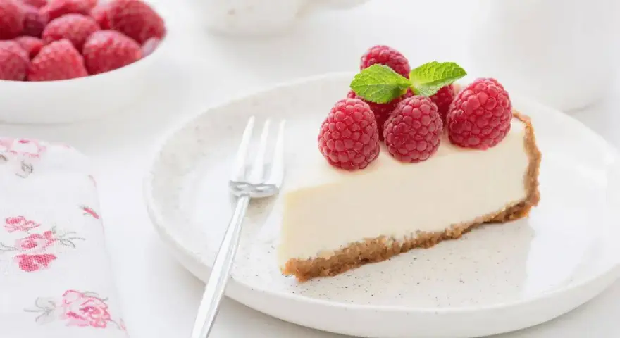 Una fetta di torta fredda allo yogurt guarnita con lamponi freschi e menta, su un piatto bianco con una forchetta.