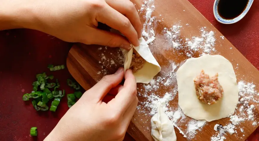 Mani che preparano il ripieno per ravioli su un tagliere di legno, con erba cipollina e salsa di soia a lato.
