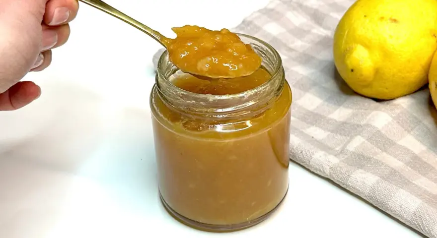 Un cucchiaio d'oro raccoglie la densa marmellata di limoni da un barattolo di vetro. Due limoni freschi riposano su un tovagliolo a quadretti.
