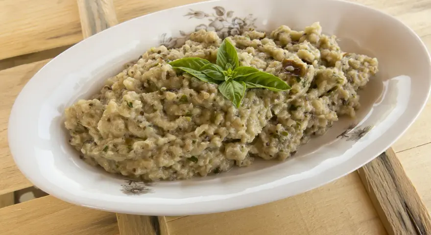 Crema di melanzane rustica, guarnita con basilico fresco, servita in un piatto ovale su un tavolo di legno grezzo.