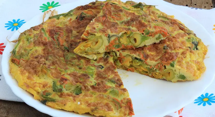 Frittata con fiori di zucca, carote e olive, tagliata a spicchi su un piatto bianco, con un tovagliolo fiorato sullo sfondo.