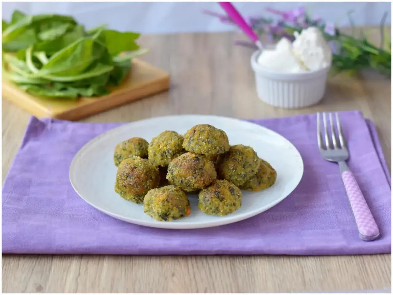 Polpette di spinaci dorate e croccanti su un piatto bianco, con spinaci freschi e una ciotolina di formaggio sullo sfondo.