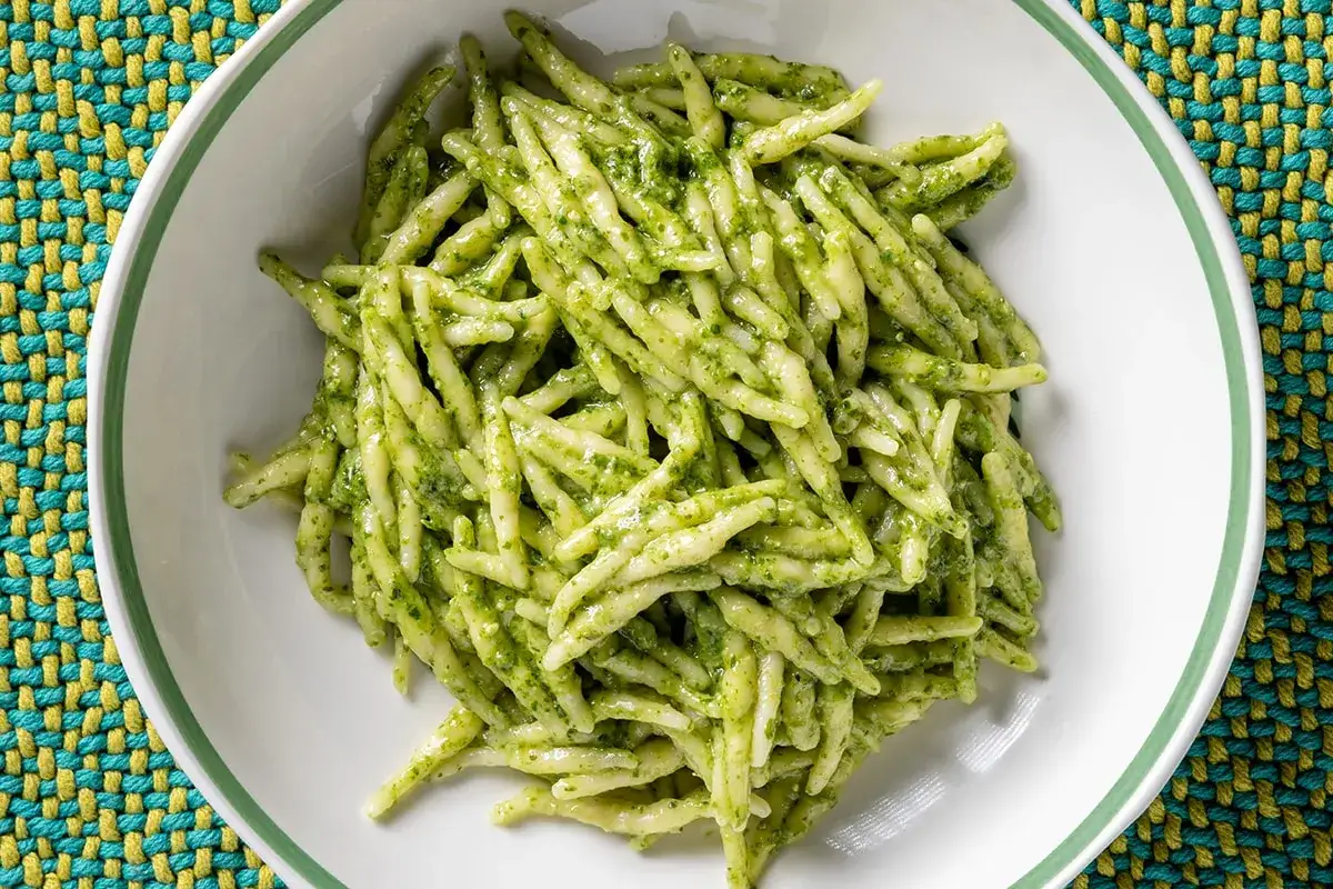 Un piatto di trofie al pesto, pasta fresca condita con un sugo verde brillante, servita in una ciotola bianca su una tovaglietta colorata.